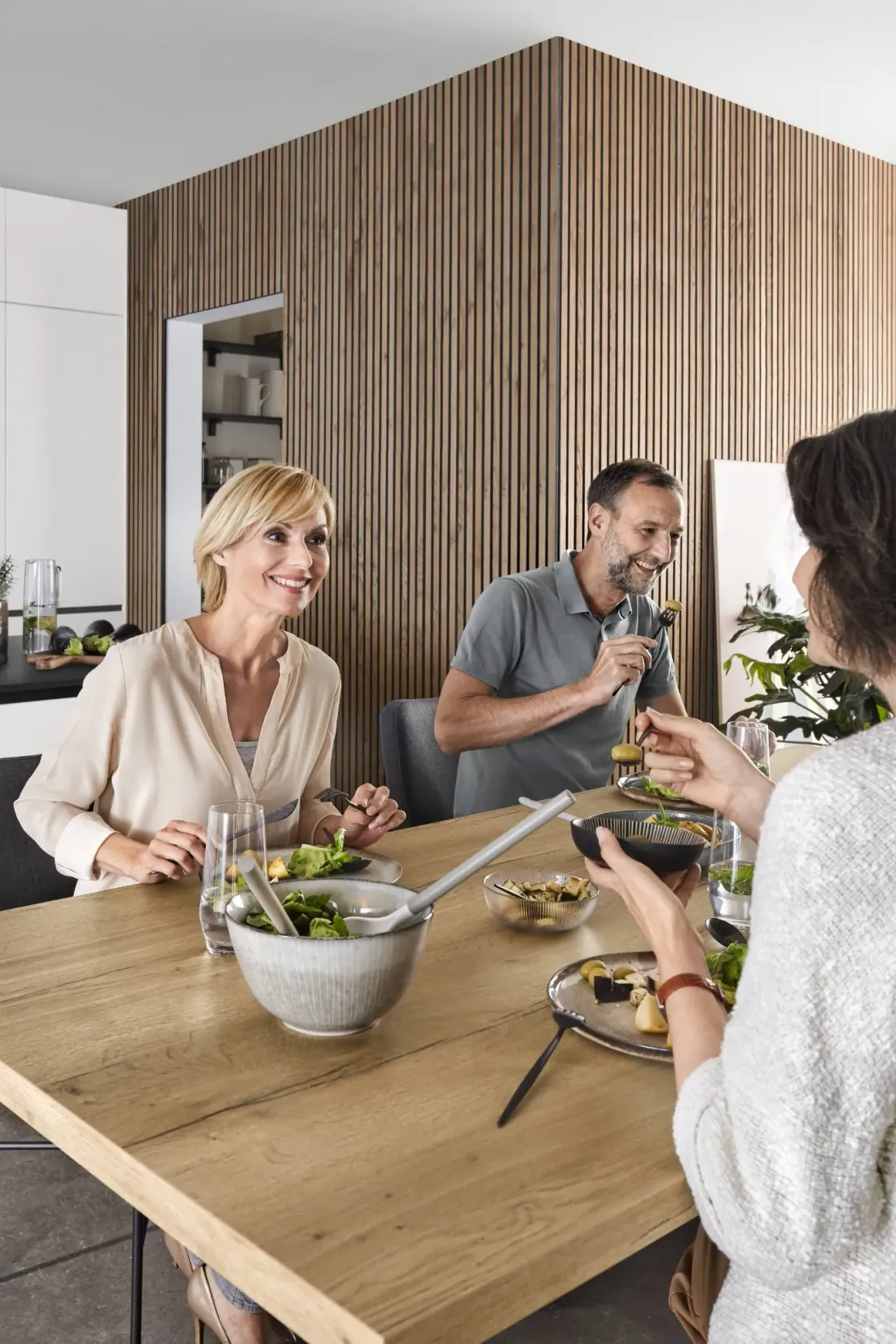 Repas partagé en famille autour de la table d’une cuisine sur mesure Cuisines ELITE, devant un mur en tasseaux de bois.